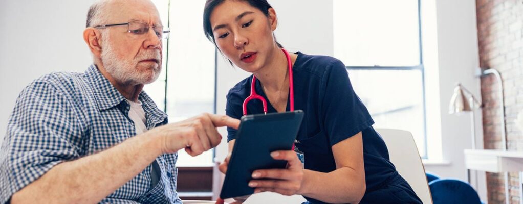 A woman with a stethoscope views a tablet with an older man who may be a patient.