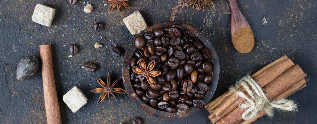 Dark roasted coffee beans in a wooden bowl surrounded by star anise, cinnamon sticks, brown sugar cubes, nutmeg, and a wooden spoon with ground cinnamon on a textured dark background.