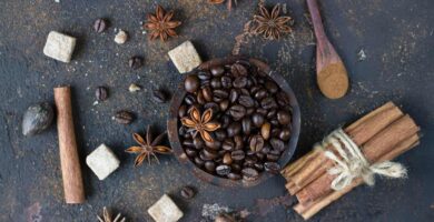 Dark roasted coffee beans in a wooden bowl surrounded by star anise, cinnamon sticks, brown sugar cubes, nutmeg, and a wooden spoon with ground cinnamon on a textured dark background.