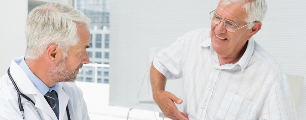 An older man in a striped shirt sits in a medical office, pointing to his side and appearing to be in discomfort, while speaking with a male doctor who is listening attentively