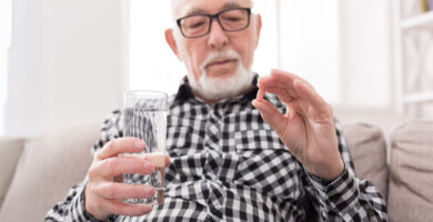 An older man holds a glass of water in one hand and a small pill in the other