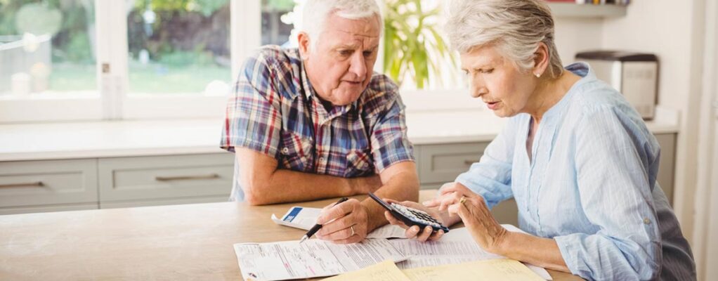 An older couple sits at their kitchen table reviewing bills with a calculator