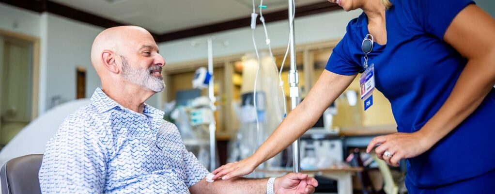 A cancer patient sits in a treatment chair receiving IV therapy while a nurse gently touches his arm in a supportive gesture