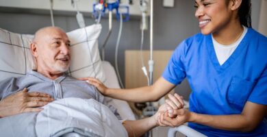 Nurse in scrubs smiling and holding the hand of an older male patient lying in a hospital bed during treatment