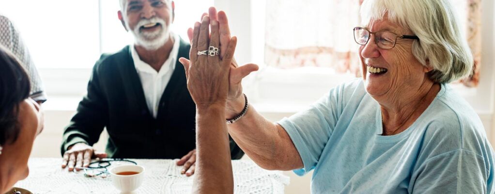 An older woman high fives another older woman sitting at a kitchen table